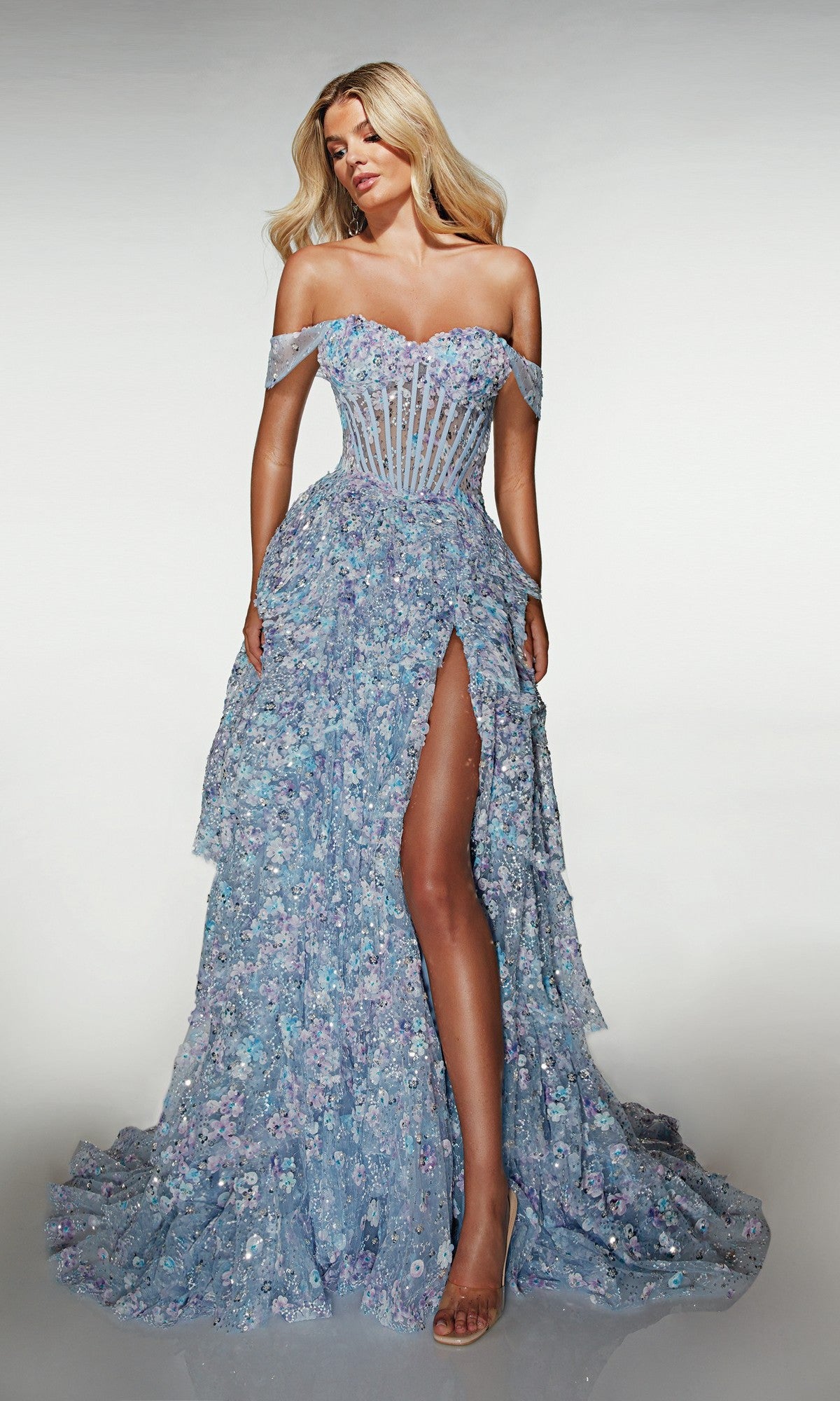 A woman poses in the Alyce 62186 long prom dress, a light blue off-the-shoulder A-line gown with a sheer corset bodice, 3D floral appliques, and a high slit, standing against a plain background.